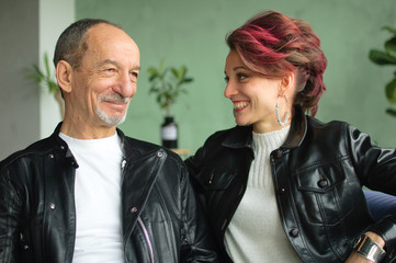 Indoor family portrait of adult daughter and senior father in loft room with houseplants. Crazy man and girl with pink hair are wearing black leather jackets in punk style and smiling