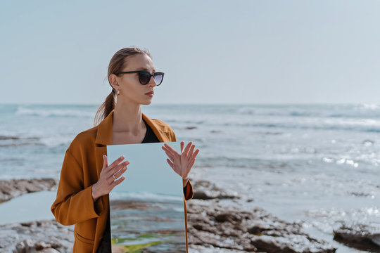 Fototapeta portrait Stylish girl in sun glasses on the sea beach holding mirror with reflection of blue sky. New vision of fashion projects concept. Dreams and illusions about travel