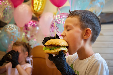 Naklejka premium The handsome little boy eating burger in black rubber gloves.
