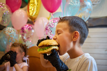 The handsome little boy eating burger in black rubber gloves.
