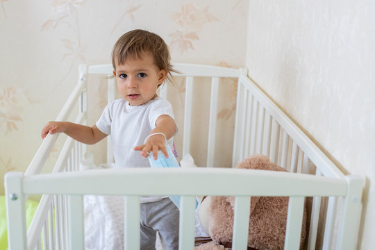 Quarantine Coronavirus Pandemic Concept. A Little Cute Baby Is In Baby Cot At Home In Safety, Looking Seriously At The Camera And Holding A Medical Mask. Copy Space