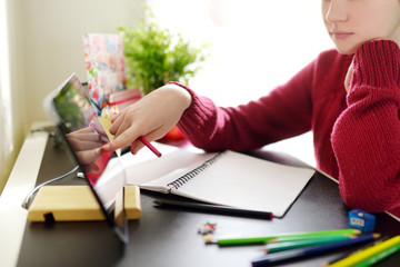 Preteen schoolgirl doing her homework with digital tablet at home. Child using gadgets to study....