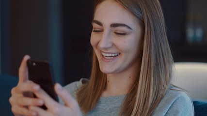 Portrait of happy pretty girl in a grey sweater with blond hair. Young woman smiling and using smartphone device or cell phone, scrolling, typing, chatting, watching video, contents, blogs