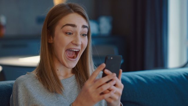 Wery happy young caucasian woman holding phone swiping in dating app feel excited finding match, overjoyed woman winner looking at smartphone screen celebrating online bid win sit on blue sofa at home