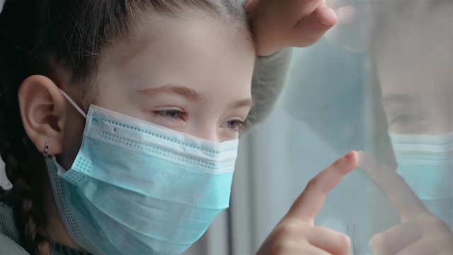 Girl in medical mask during quarantine looks sadly at her reflection in window. Stop COVID-19 infection