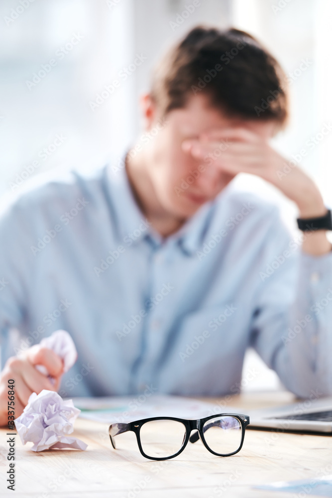 Poster tired man being puzzled with paperwork sitting at desk with crumpled paper in hand, focus on eyeglas