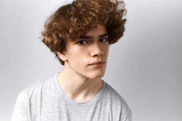 Pretty boy with messy voluminous hairstyle and clean shaven face looking at camera with curious facial expression. Attractive young man in gray casual t-shirt being interested in communication