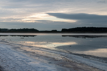Wald mit Ufer spiegelt sich im Wasser an einem ruhigen Wintertag, im Vordergrund das vereiste Ufer, dramatischer Wolkernhimmel