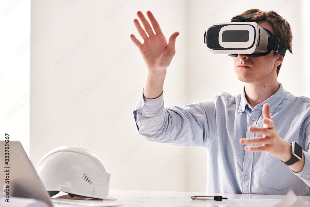 Aufkleber Concentrated young engineer sitting at table and twisting hands while using virtual reality device to make 3D building project