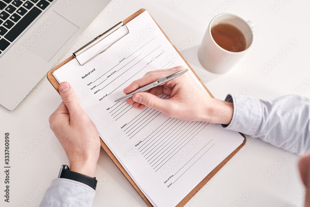 Poster Above view of unrecognizable manager sitting at table with mug and laptop and filling personal information in medical card