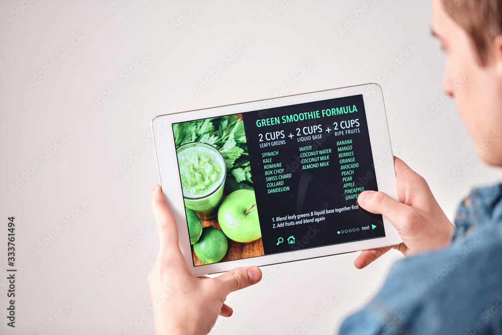 Poster over shoulder view of young man leading healthy lifestyle using tablet while reading green smoothie 