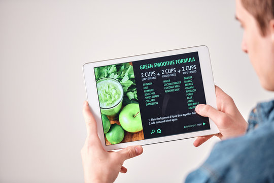 Over shoulder view of young man leading healthy lifestyle using tablet while reading green smoothie recipe on website