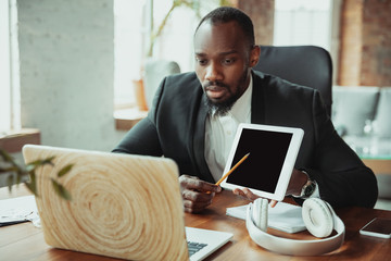 Businessman or student working from home being isolated or keep quarantine 'cause of coronavirus. African-american man using laptop, tablet and headphones. Online conference, lesson, remote office.