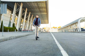 Young handsome man riding the e-scooter on the road near the airport on the sunset. Trendy urban transportation on modern electric scooter. Eco friendly mobility concept.