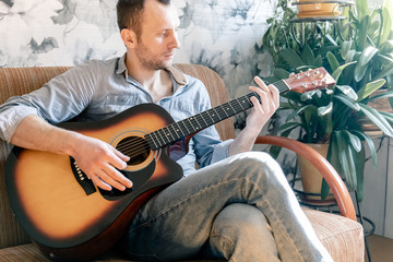 young man playing an acoustic guitar while sitting on a sofa in an apartment