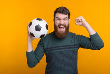 Excited bearded man holding a ball in his hand while screamin and making winner gesture on yellow background.