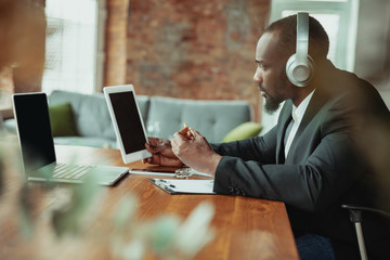 Businessman or student working from home being isolated or keep quarantine 'cause of coronavirus. African-american man using laptop, tablet and headphones. Online conference, lesson, remote office.