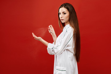 Young woman in medical uniform sprays a sanitizer on her hand, isolated on a red background. Female doctor use sanitizer spray for disinfecting her hands. Concept of healthcare and medicine