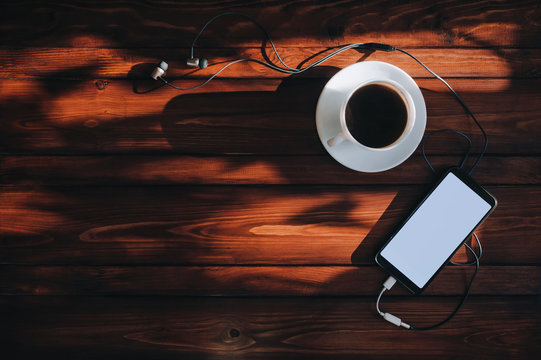 Black smartphone with white screen, dark headphone and cup of coffee lies on a brown wooden background mahogany with spots of sunlight. Top view, copy space, mockup. Morning breakfast, music concept. - Powered by Adobe