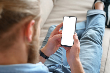 Young male tech user relaxing on sofa holding smartphone mock up blank white screen. Man customer...