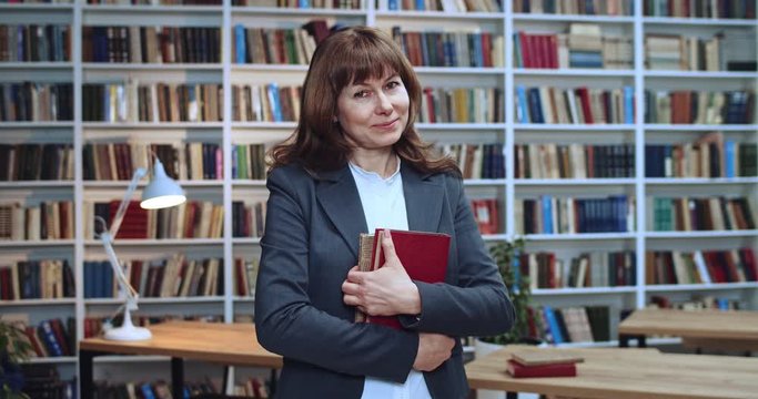 Close-up portrait of midle aged woman standing in library and hilding books at hands next to book rack and looking at camera.