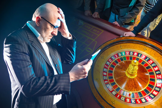 Losing In An Online Casino. A Man With A Tablet In His Hands On The Background Of A Roulette Wheel. Gambling For Money. Losing In An Online Casino. Losses From Online Games.