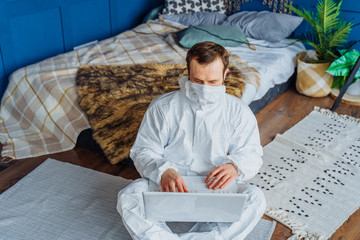Portrait of caucasian man in white viral protective costume and face mask sitting near the bed and working from home on laptop because of coronavirus. Isolated at home due to quarantine covid-19