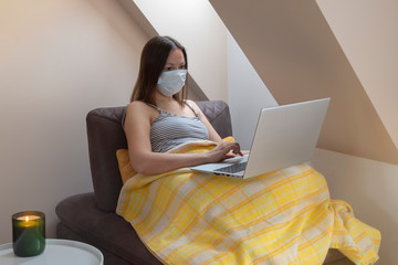 Young woman wearing medical mask on quarantine holding laptop sitting on sofa by the window