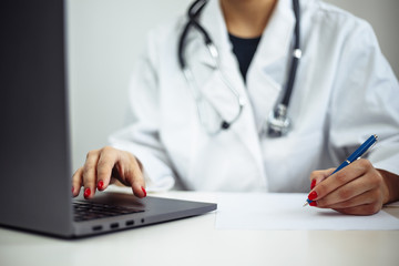 Closeup of a woman doctor wearing white medical gown and blue gloves, stethoscope. Female nurse sits at the desk with laptop and papers diagnosis. Coronavirus, covid-19, sickness prevention concept.