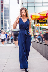 Fototapeta premium Young Woman Street Fashion in New York City. Young Eastern European Woman wearing blue sleeveless, v neck jumpsuit, black leather shoes, walking on busy street in middletown of Manhattan, relaxing..