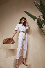 Beautiful woman fashion model brunette hair tanned skin wear white overalls button suit sandals high heels accessory bag clothes style journey safari summer collection plant flowerpot wall stairs. - obrazy, fototapety, plakaty