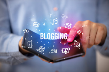 Businessman holding a foldable smartphone with BLOGGING inscription, social media concept