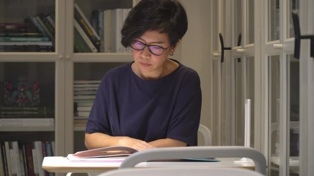 Stylish, short hair middle aged Asian woman sit alone in relaxing, calm library cafe, flip through book to study for her work, surrounded by wall bookshelves. Social distancing and introvert concept.