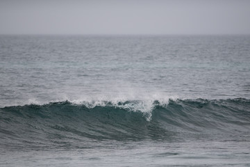 The start to a long wave with a spray coming over the top of the curl. It's a grey gloomy day. The sky and water are pale grey in color. The water is in both the foreground and background.