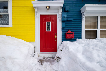 Red door of a building with blue and yellow wooden clapboard walls. The house has a red metal...