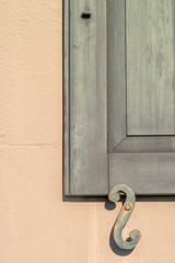 Close up of an antique shutter latch holding a green wooden shutter open against a beige building. 