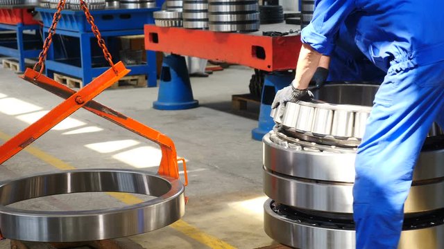 Back view of male worker working in busy bearing factory warehouse, wearing blue uniform, checking and operating a crane, lifting metal parts of huge round detail with crane-beam. 4 k footage