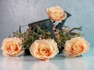 4 peach roses and a floral pink and green wreath with a rustic metal watering can with a white counter and a plaster wall.  Simple, soft, country home decor.