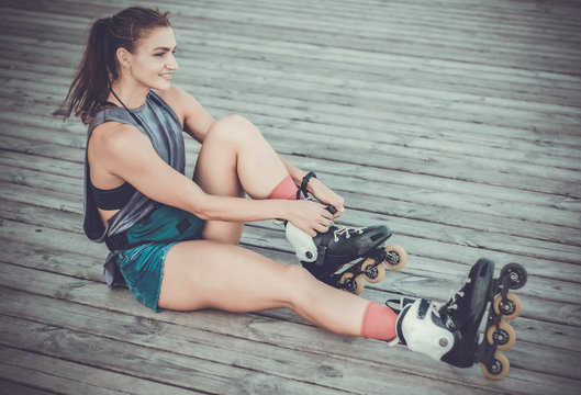 Young happy woman roller skater sitting on wooden boards and trying on roller skates outdoors. Sport lifestyle