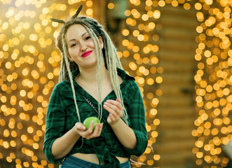 An informal young woman with a dreadlocks hairstyle and fashionable summer clothes holds a ripe green apple against the background of garlands