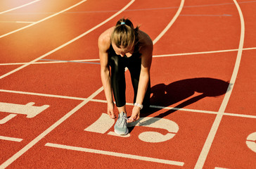 Young sprinter woman in sportswear tying shoelaces before a race on a red-coated stadium track at sunny bright day