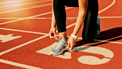 Young sprinter woman in sportswear tying shoelaces before a race on a red-coated stadium track at sunny bright day
