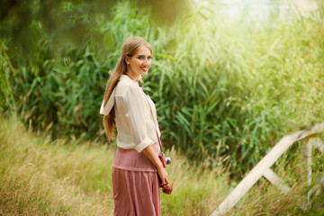 Young smiling woman in vintage style clothes with retro camera against the background of reeds on the lake