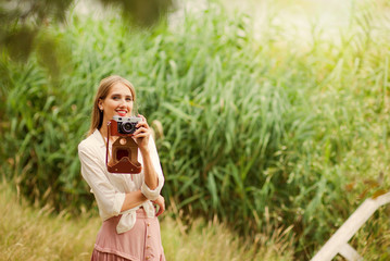 Young smiling woman in vintage style clothes with retro camera against the background of reeds on the lake