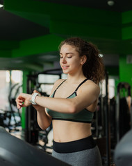 Fototapeta premium Fit woman in sportswear runs on a treadmill and looks at her watch in the gym. The concept of a healthy lifestyle, warming up, fitness, weight loss.