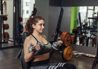 Fototapeta premium Young fit woman doing exercise lifting dumbbell with hands while sitting on a bench in the gym