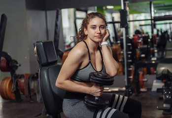 Tired young fit woman holding dumbbells and resting while sitting on a bench in the gym