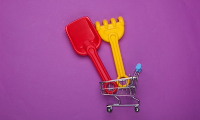 Children's rake and shovel for a sandbox or beach in shopping trolley on purple studio background....