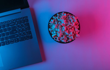 Laptop and bowl of popcorn with blue pink neon light. Leisure and entertainment concept. Top view