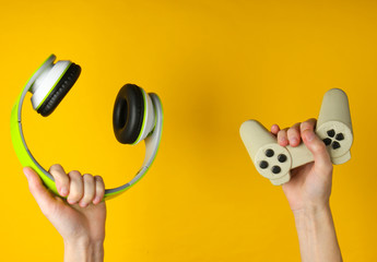 Hands hold stereo headphones and gamepad on yellow background. Gaming, recreation and entertainment